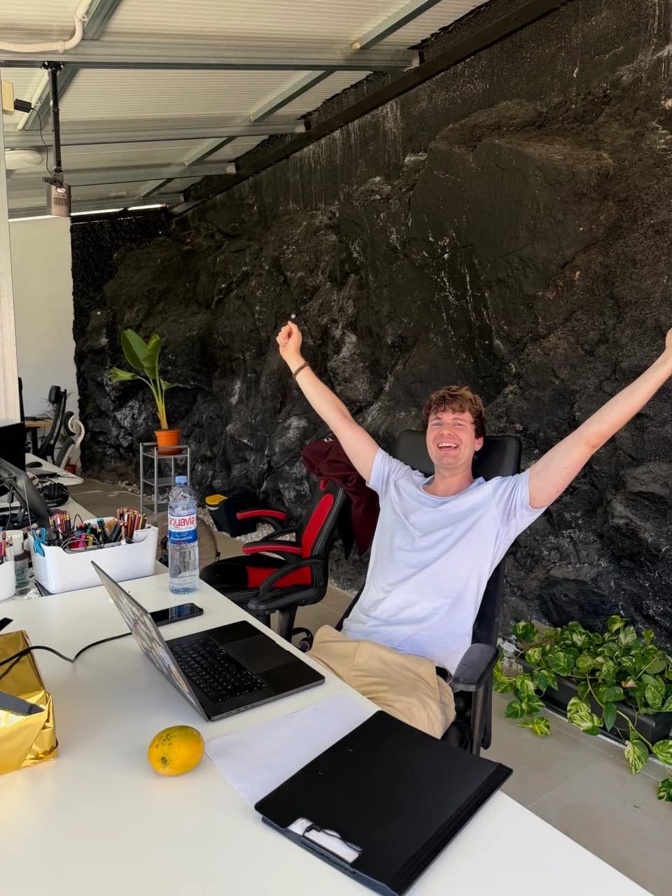 Smiling remote worker raising both arms at a desk with a laptop beside the volcanic rock wall in Blue Paradise’s coworking