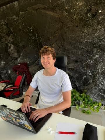 Jakob smiling while working on his laptop at a desk beside a stone wall.