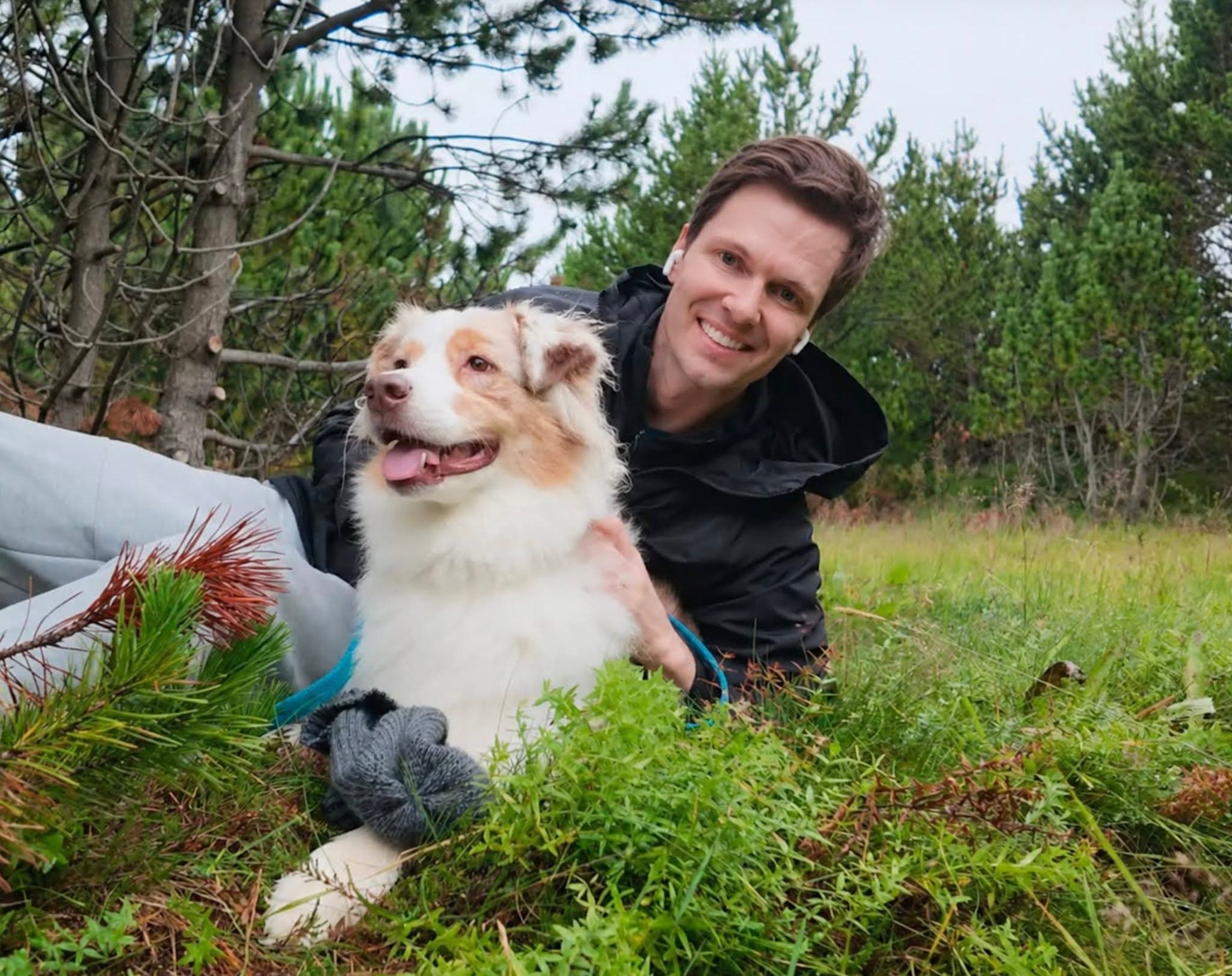 Jakob sorride accanto a un cane bianco e marrone in una pineta dal suolo erboso.